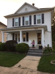 Two story house with a front porch. There are three steps leading up to the porch with blue brick steps that are lined up vertically at the front of the step. There is an adorable 3 year old girl standing proudly at the top of the steps as if she had done them herself.